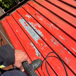 DIZ POL Podnośnik koszowy, Wycinka drzew, Mycie ciśnieniowe - Czerwony dach z blachy trapezowej, częściowo zdarta farba, widok z góry podczas mycia. W tle widać zieleń i fragment posesji.