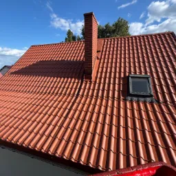 Czerwony dach z dachówki ceramicznej z ceglanym kominem i oknem dachowym, widziany z góry, na tle błękitnego nieba z chmurami. Widoczne drzewa w tle.