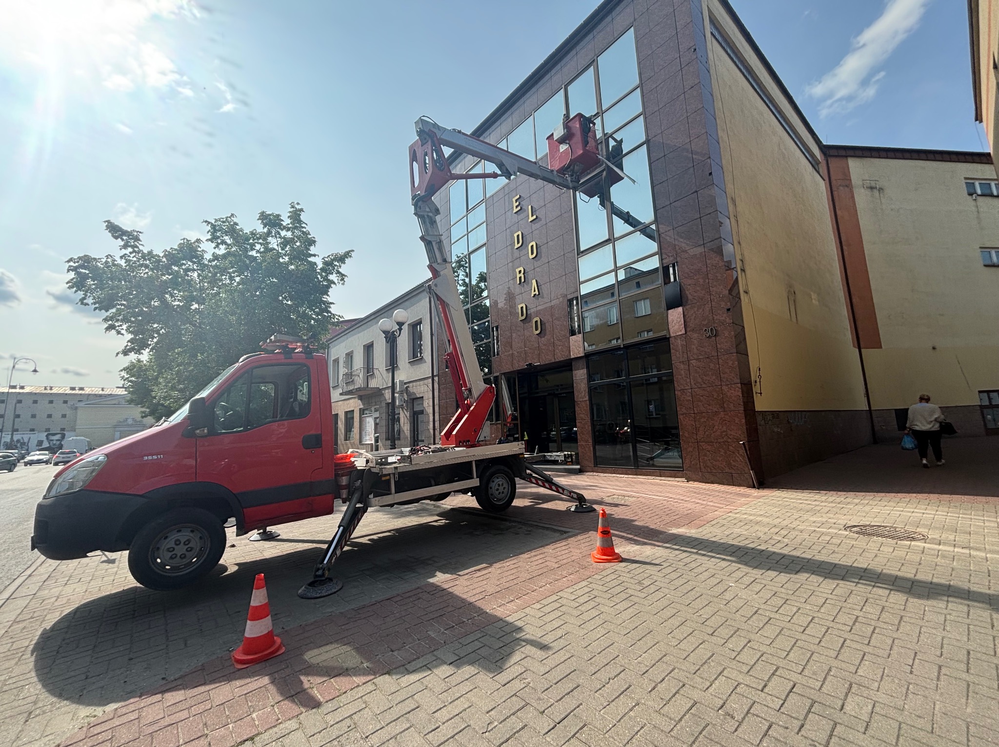 Czerwony podnośnik koszowy czyści szklaną fasadę budynku Eldorado, z widocznymi odbiciami, na tle błękitnego nieba. Pomarańczowe pachołki ostrzegawcze na chodniku.