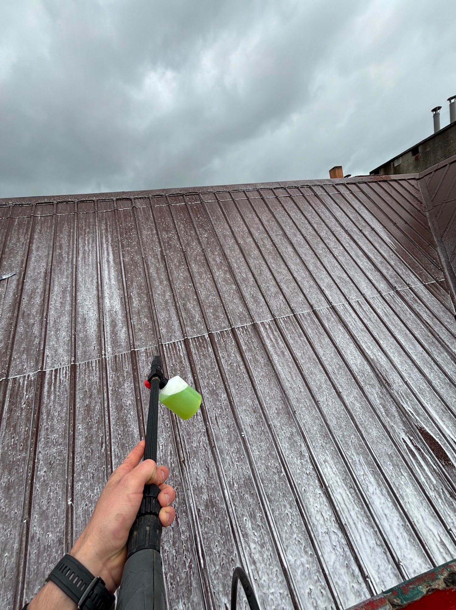 Czyszczenie brązowego dachu z blachy za pomocą myjki ciśnieniowej z zielonym detergentem, widok z dołu na pochmurne niebo.