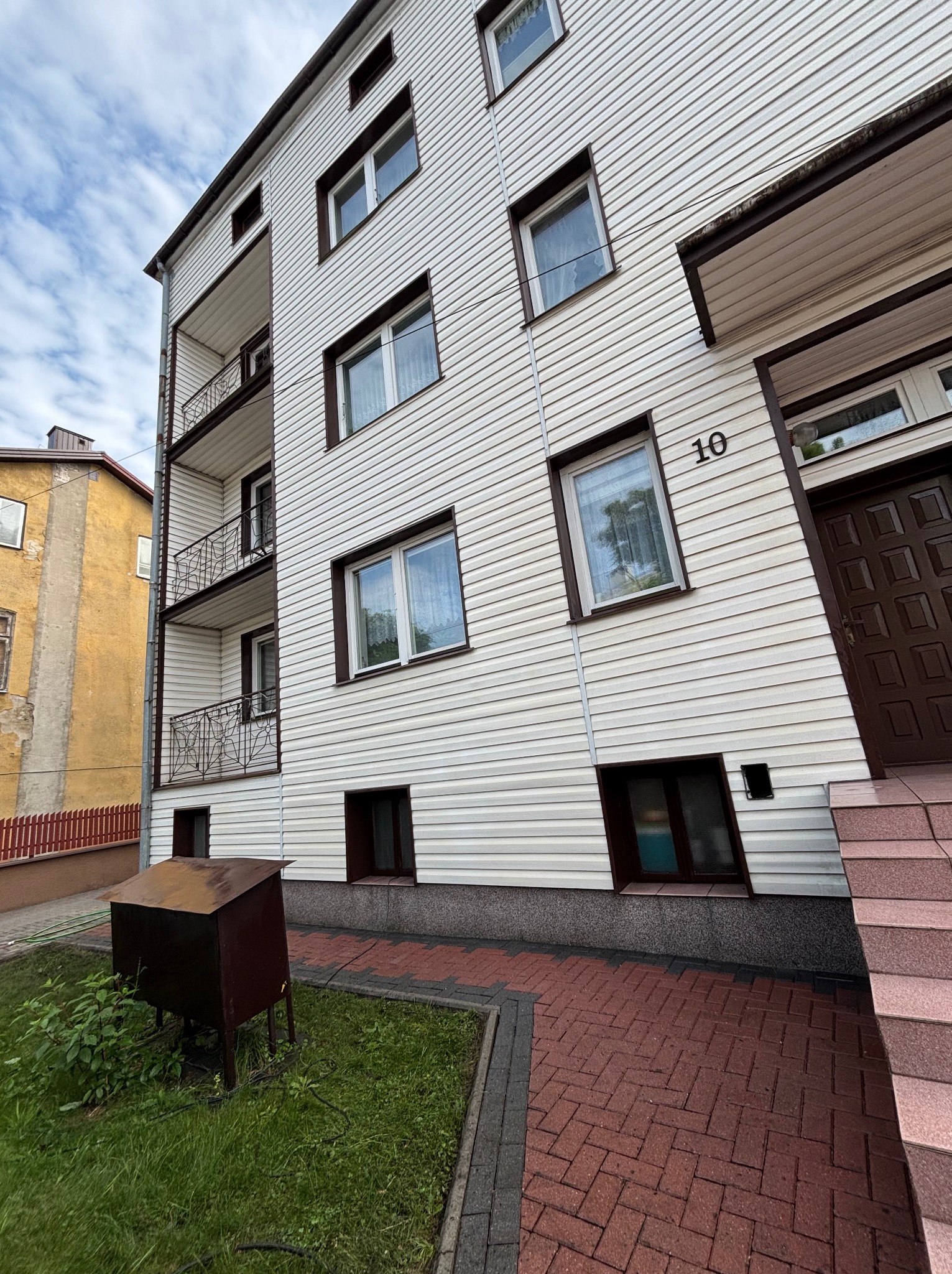 Trzypiętrowy budynek z białym sidingiem, brązowymi ramami okiennymi i balkonami. Widoczny numer domu '10' i fragment chodnika z kostki brukowej.