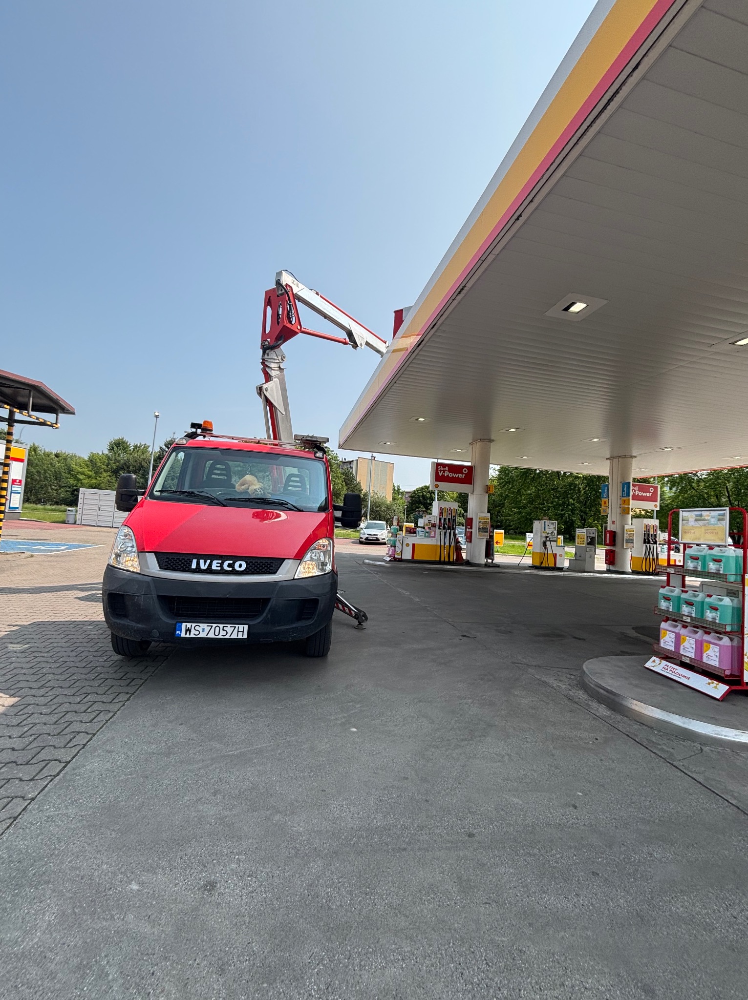 Czerwony samochód Iveco z podnośnikiem koszowym przy stacji benzynowej Shell. Widoczny wysięgnik i elementy konstrukcyjne stacji. Słoneczny dzień.