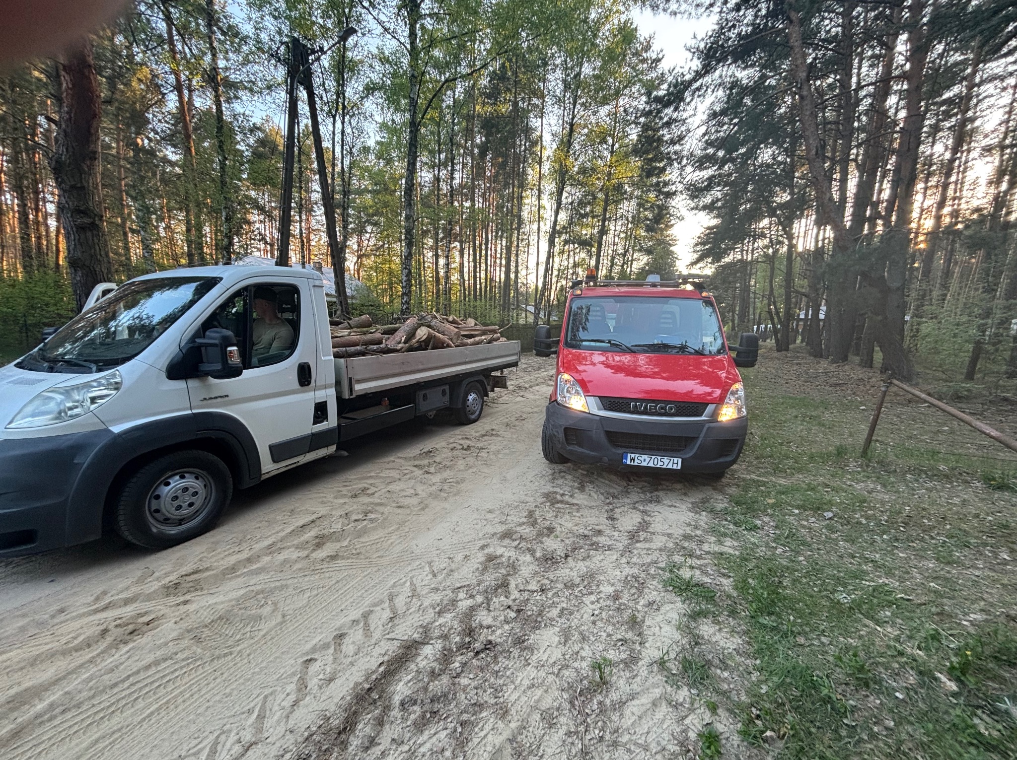 Dwa samochody dostawcze na leśnej drodze, jeden z drewnem, sugerujące wycinkę lub transport drewna. Widoczne drzewa w tle.