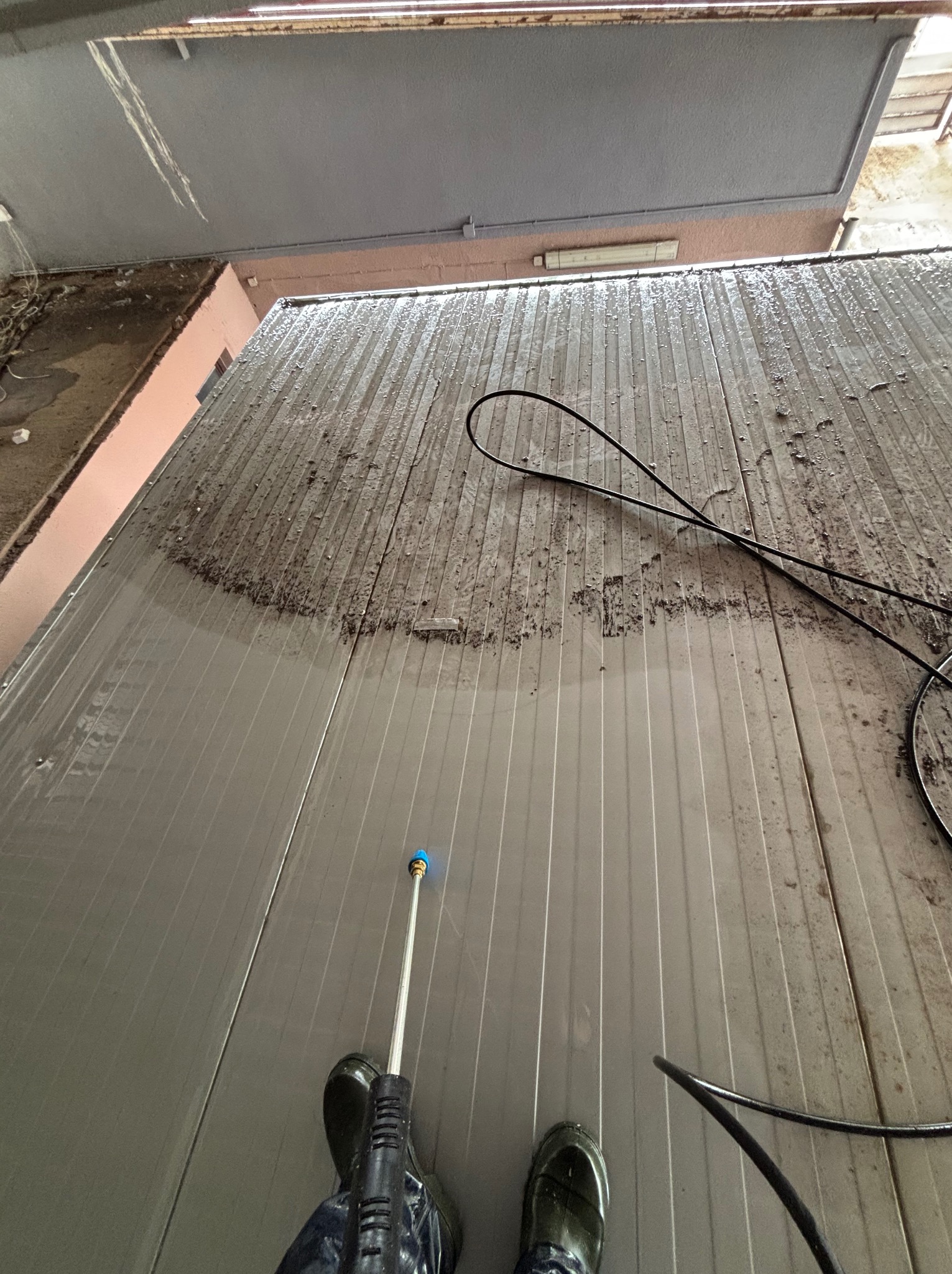 Częściowo umyta, brudna, metalowa powierzchnia dachu z widocznym operatorem myjki ciśnieniowej w gumowych butach. Widok z góry, z bliska.