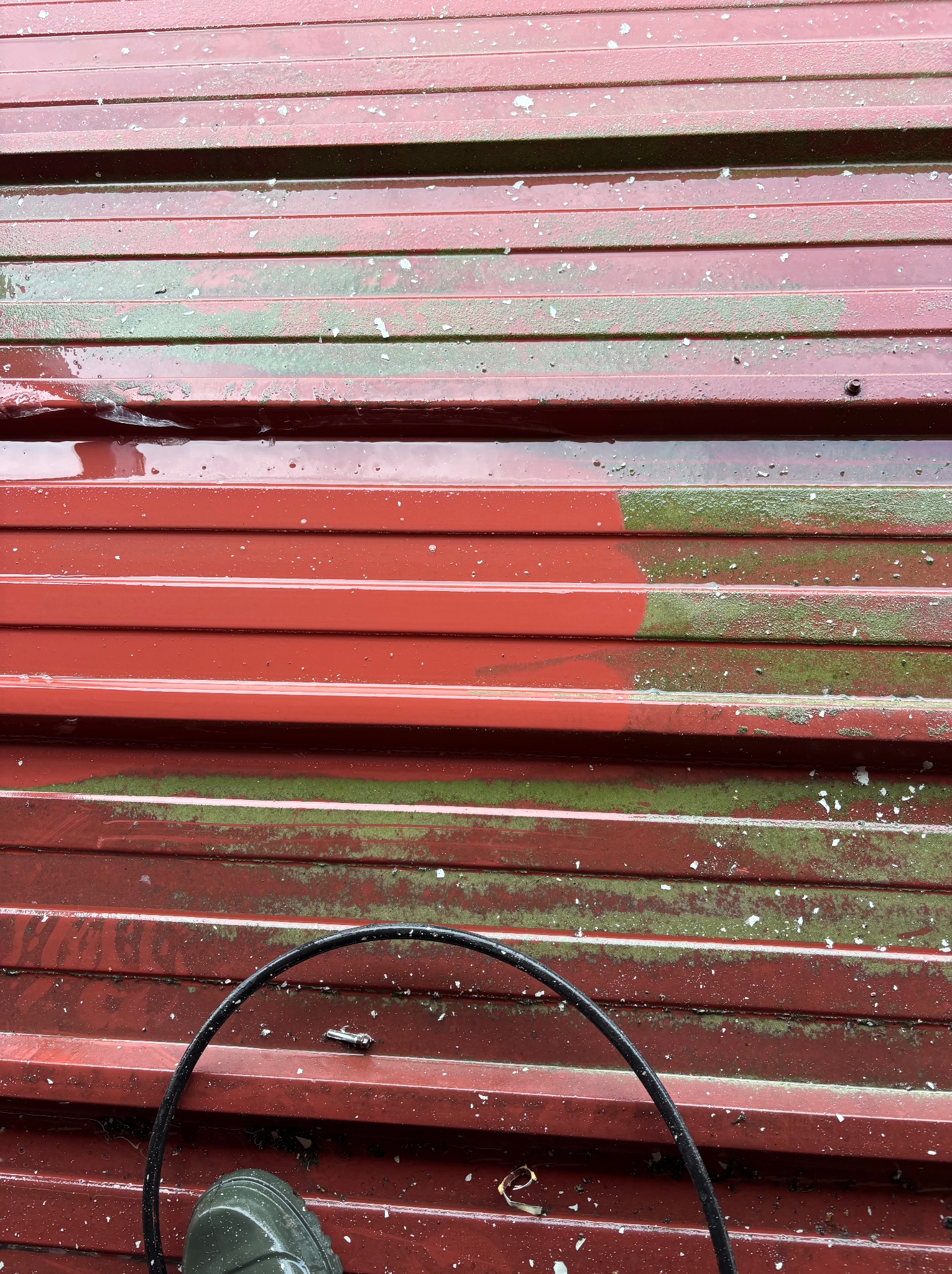 Czerwony dach z blachy trapezowej, częściowo umyty, z widocznymi śladami mchu i resztkami białego osadu. Na dachu wąż ciśnieniowy i but roboczy.