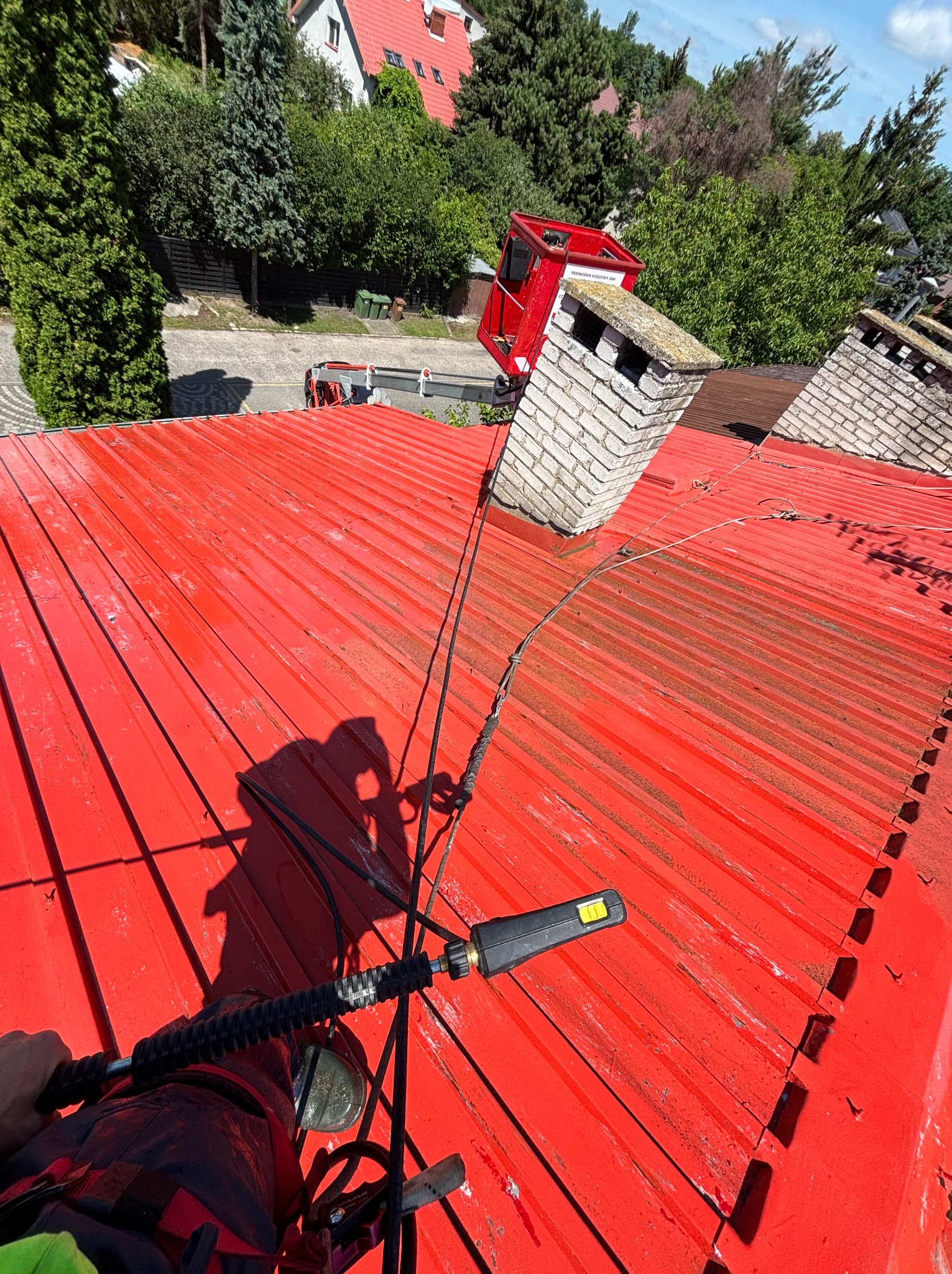 Czerwony dach z blachy trapezowej, czyszczony lancą ciśnieniową. W tle komin i podnośnik koszowy. Widoczny cień osoby wykonującej prace na wysokości.