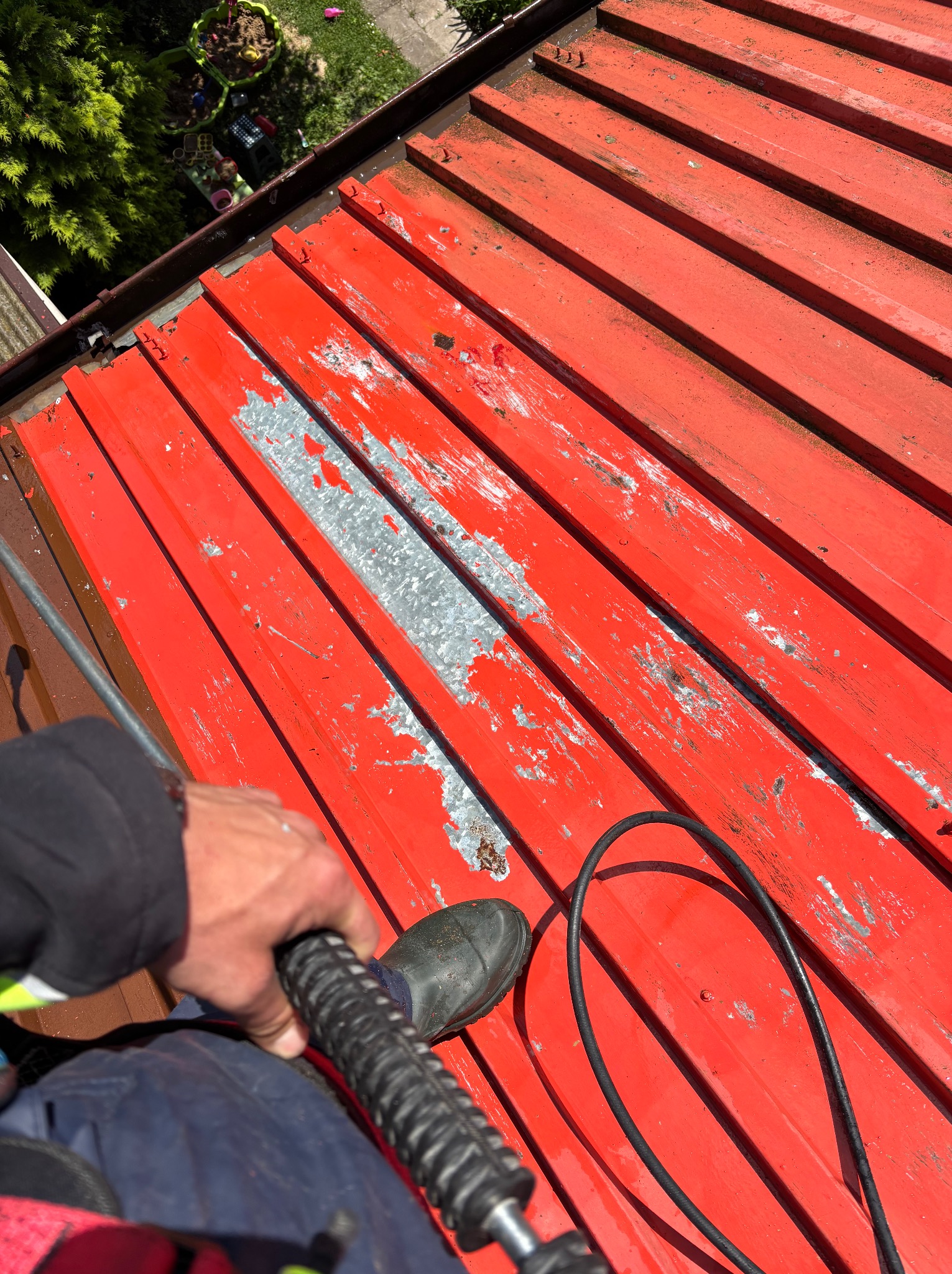 Czerwony dach z blachy trapezowej, częściowo zdarta farba, widok z góry podczas mycia. W tle widać zieleń i fragment posesji.