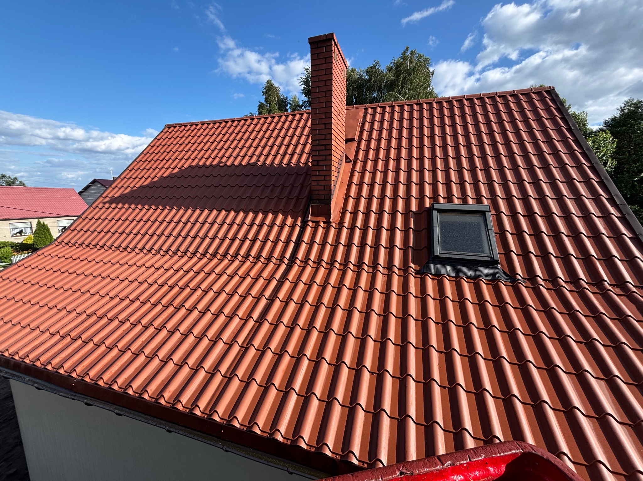 Czerwony dach z dachówki ceramicznej z ceglanym kominem i oknem dachowym, widziany z góry, na tle błękitnego nieba z chmurami. Widoczne drzewa w tle.