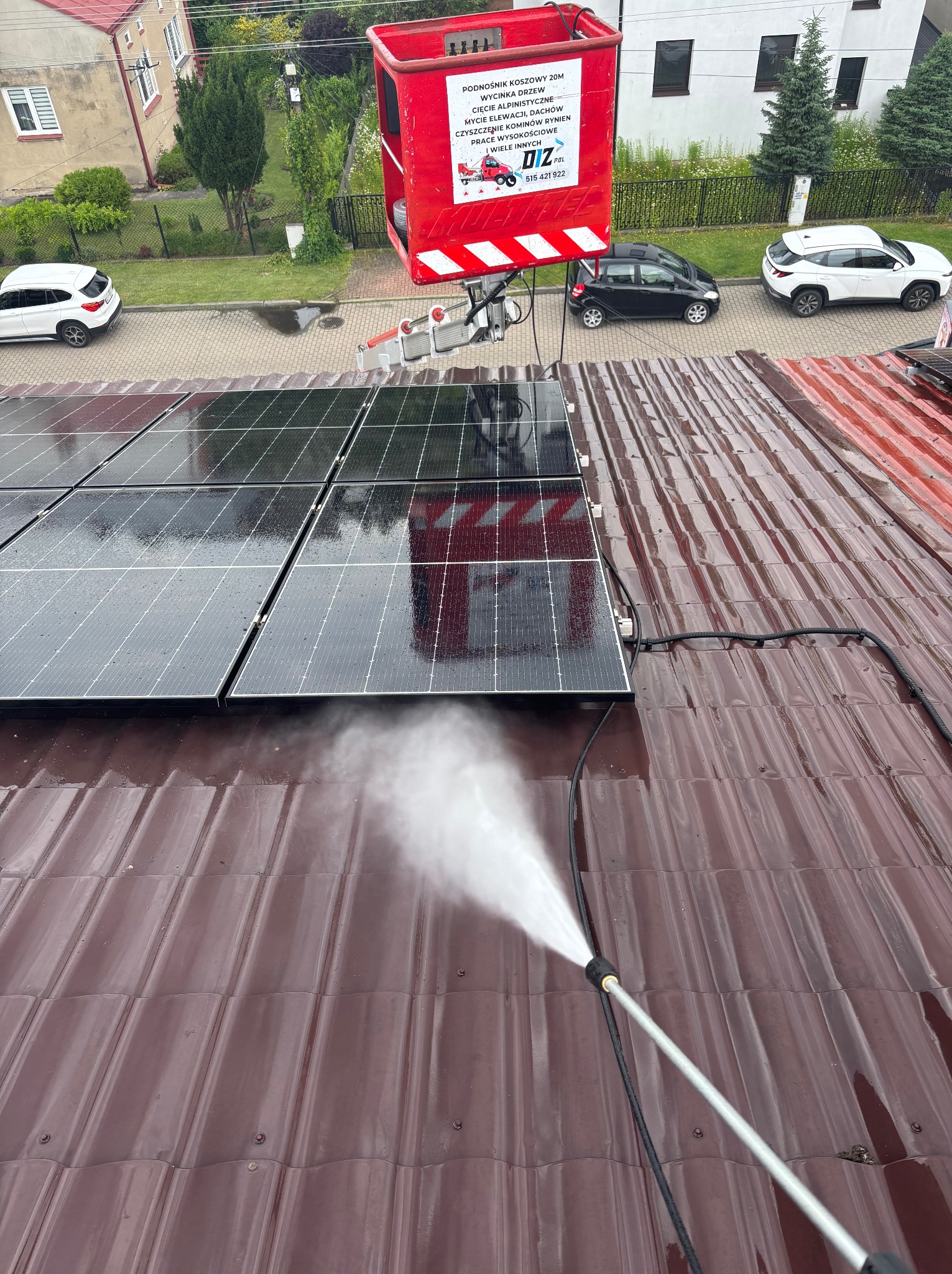 Czyszczenie paneli słonecznych na brązowym dachu za pomocą myjki ciśnieniowej z podnośnika koszowego, widok z góry. W tle zabudowa mieszkalna i zaparkowane auta.