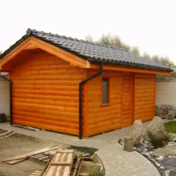 Drewniany domek narzędziowy z ciemną dachówką, rynną i małym oknem, otoczony kostką brukową i elementami dekoracyjnymi z kamienia.