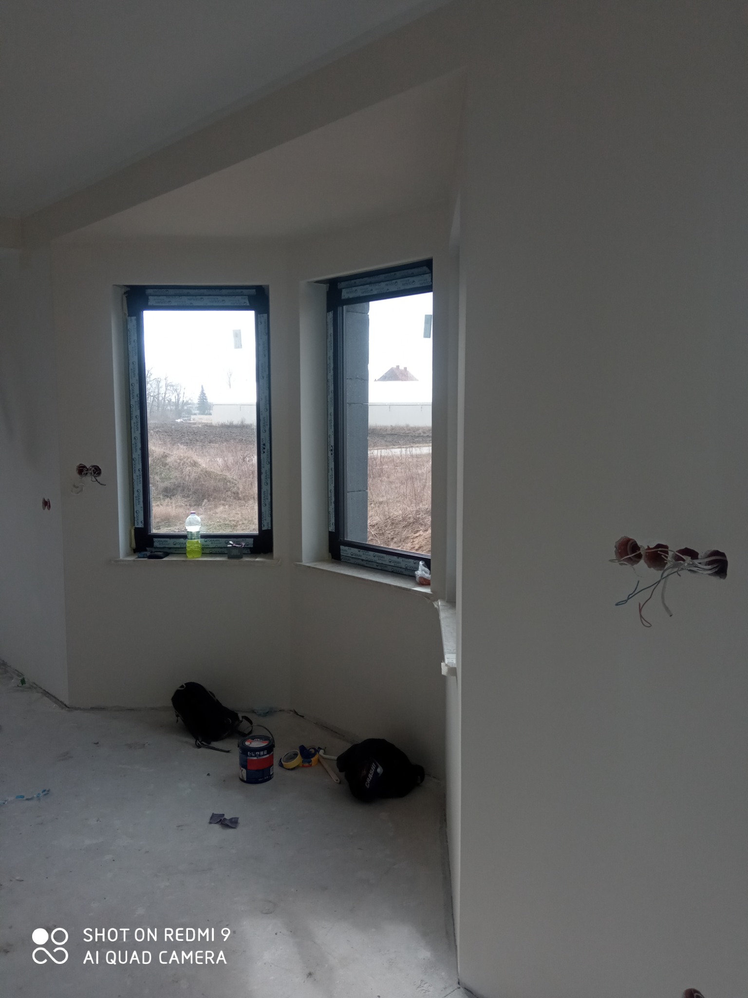 Wnętrze w stanie deweloperskim po nałożeniu gładzi, widoczne puszki elektryczne bez osprzętu, okna z widokiem na zewnątrz, na podłodze wiadro z farbą, torby i akcesoria malarskie.