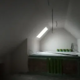 Poddasze po nałożeniu białych tynków, widoczne okno dachowe i wystające przewody elektryczne. Przy ścianie ustawione zielone pojemniki.