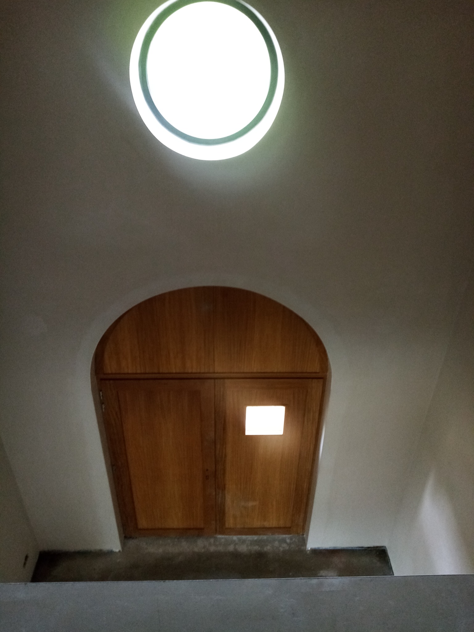 Wiatrołap z drewnianymi drzwiami o zaokrąglonym kształcie, oświetlony okrągłym świetlikiem w suficie i małym okienkiem w drzwiach.