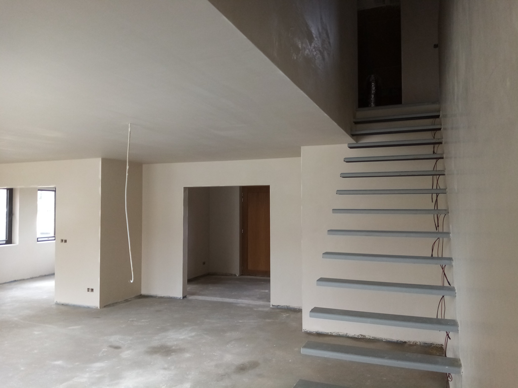 Wnętrze w stanie deweloperskim po nałożeniu gładzi, widoczne fragmenty instalacji elektrycznej przy schodach, surowy beton na podłodze.