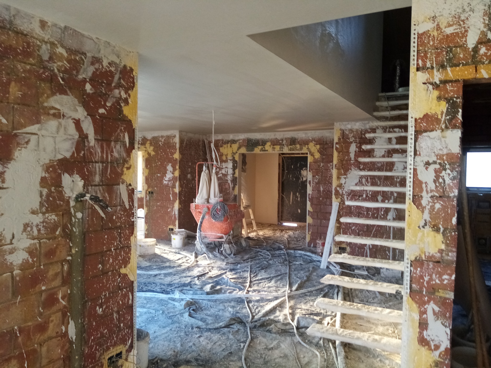 Wnętrze w trakcie remontu: ściany z fragmentami starego tynku, zabezpieczona folią podłoga, maszyna do tynkowania w centralnej części pomieszczenia, widoczne schody w tle.