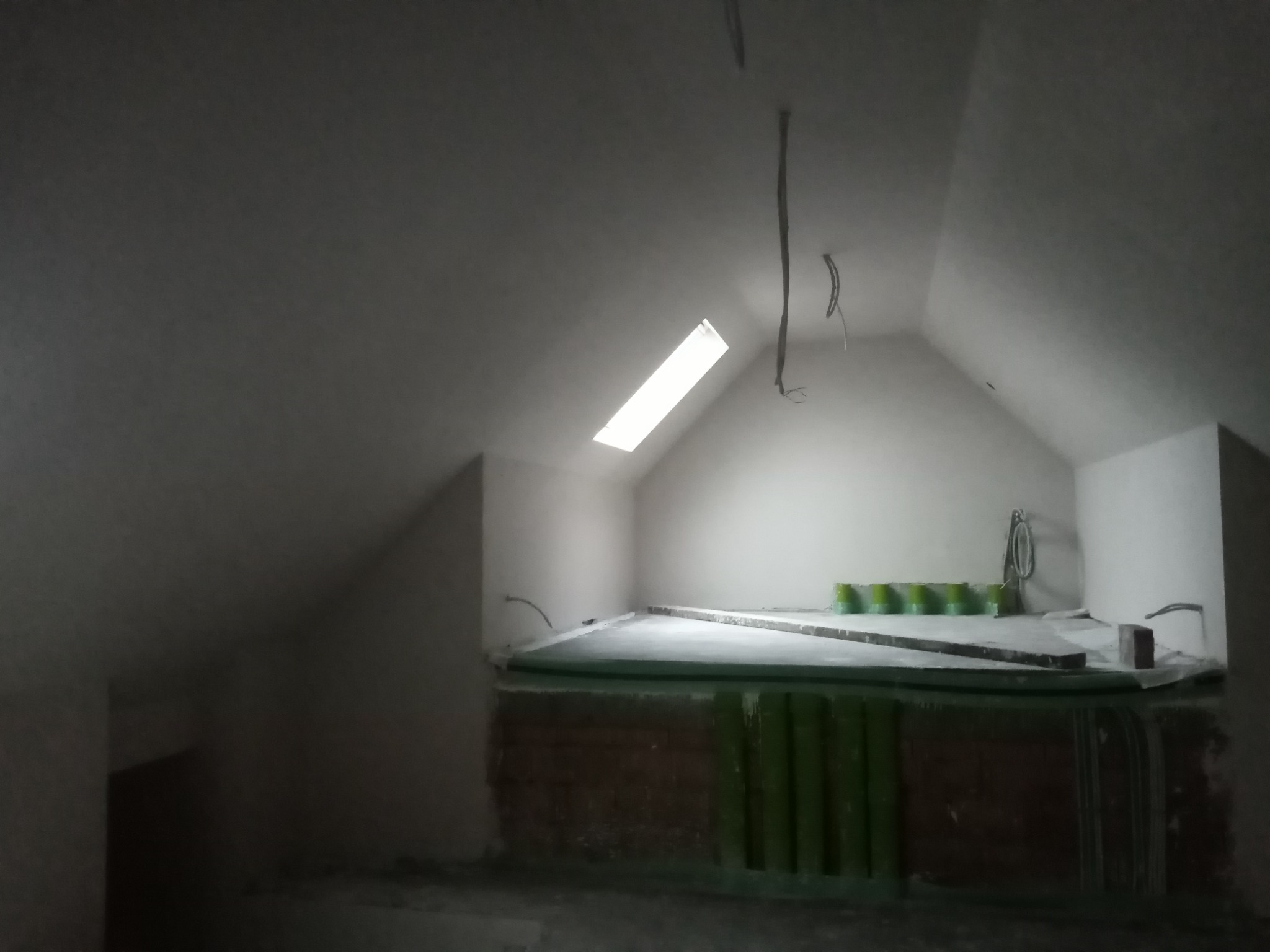 Poddasze po nałożeniu białych tynków, widoczne okno dachowe i wystające przewody elektryczne. Przy ścianie ustawione zielone pojemniki.