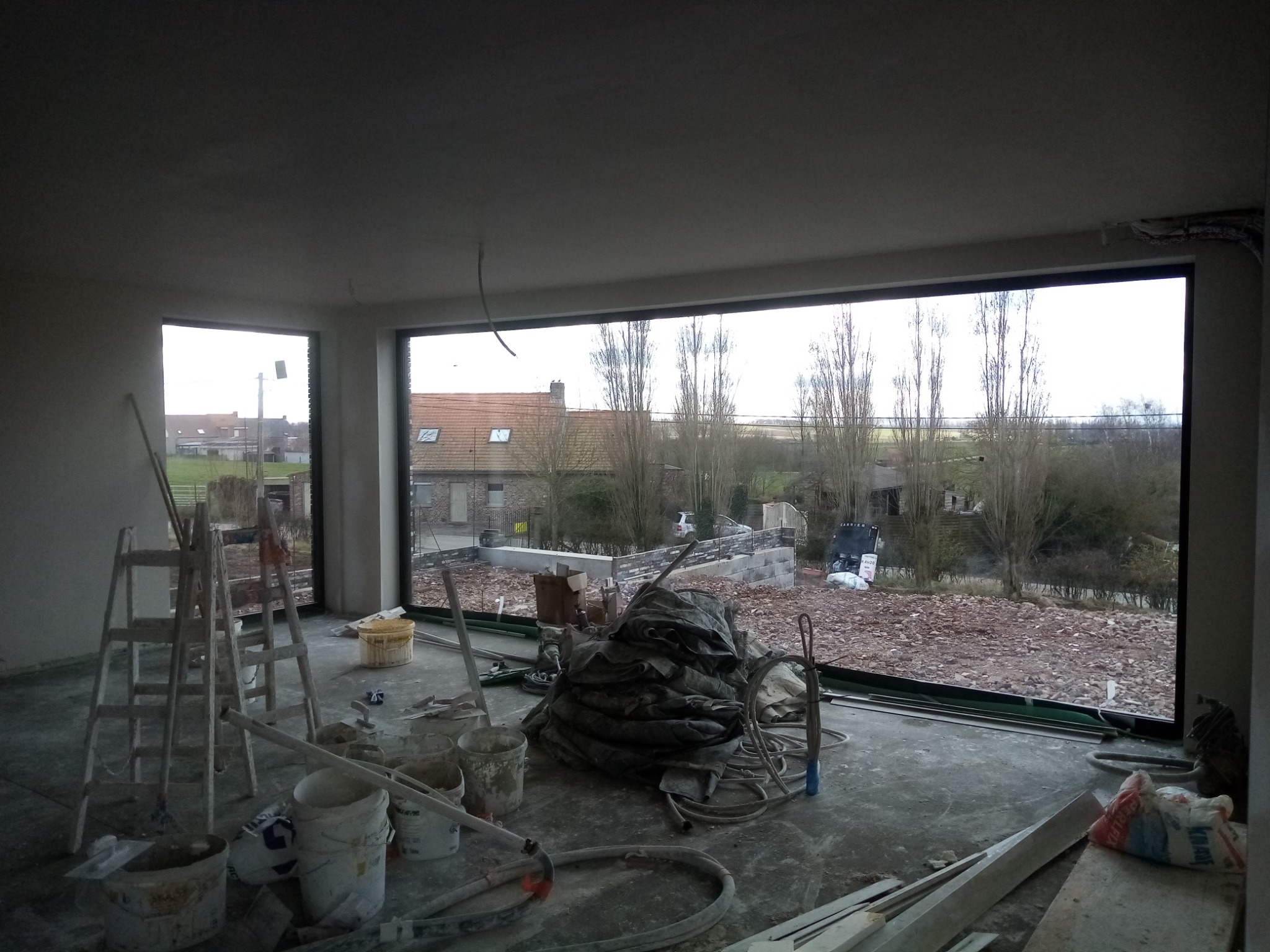 Wnętrze w trakcie prac wykończeniowych: widoczne ściany po nałożeniu gładzi, drabina, wiadra z zaprawą i okno z widokiem na zewnątrz.