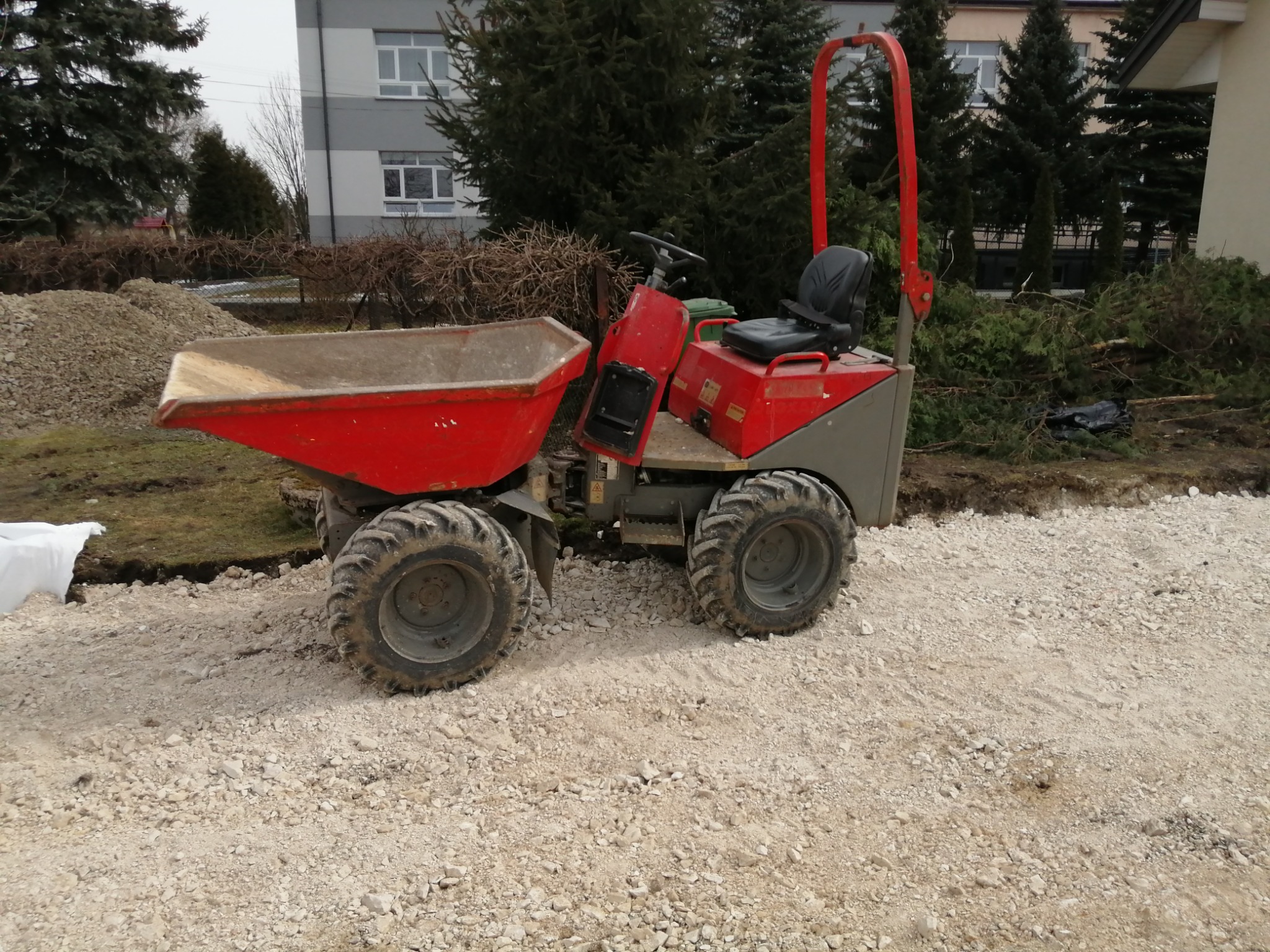 Czerwona mini wywrotka na drodze z tłucznia, w tle budynek i drzewa iglaste.