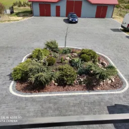 Szara kostka brukowa ułożona na posesji z wyspą zieleni na środku, widok z góry, w tle budynki gospodarcze i zaparkowane samochody.