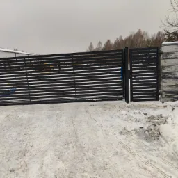 Nowoczesna, pozioma brama wjazdowa z czarnych, płaskich profili, osadzona na słupkach z szarego, łupanego kamienia, na tle zimowego krajobrazu z zaśnieżonym podjazdem i drzewami.