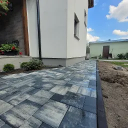 Ułożona ścieżka z kostki brukowej w odcieniach szarości i błękitu, obrzeża z czarnego kamienia, widoczny drenaż liniowy, tło stanowi budynek z białej elewacji i fragment ogrodu.