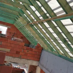 Wnętrze w trakcie budowy domu murowanego: widok na zieloną konstrukcję dachu z drewnianych belek i membraną dachową, ściany z czerwonej cegły ceramicznej porotherm, oraz fragment betonowego...