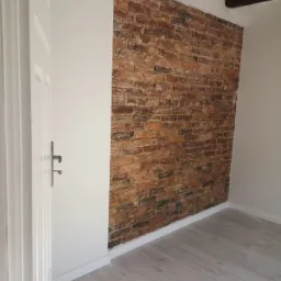 Ściana z cegły dekoracyjnej w odnowionym wnętrzu, widoczne drewniane belki stropowe, białe ściany i panele podłogowe w odcieniach szarości, białe listwy przypodłogowe oraz fragment białych drzwi...