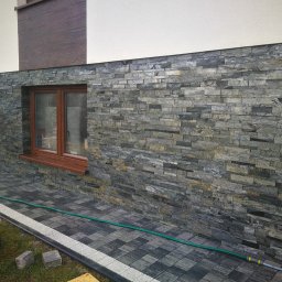 Pachota Paweł PROGRESS Usługi Remontowo Budowlane - Elewacja budynku wykończona szarą, łupkową okładziną kamienną w wąskich pasach, z widocznym oknem z brązową ramą, dopasowana kolorystycznie do kostki brukowej i zarośniętym trawnikiem u podstawy.