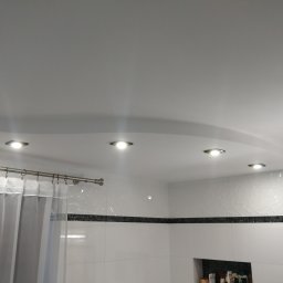 Pachota Paweł PROGRESS Usługi Remontowo Budowlane - Nowoczesna łazienka z białymi, błyszczącymi płytkami, czarnym pasem dekoracyjnym i podwieszanym sufitem z czterema punktowymi lampami LED, widoczna zasłona prysznicowa.