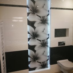 Pachota Paweł PROGRESS Usługi Remontowo Budowlane - Wykończona łazienka z biało-czarnymi płytkami, podświetlaną wnęką z motywem kwiatowym, wiszącą toaletą i grzejnikiem drabinkowym, drewnopodobna podłoga.