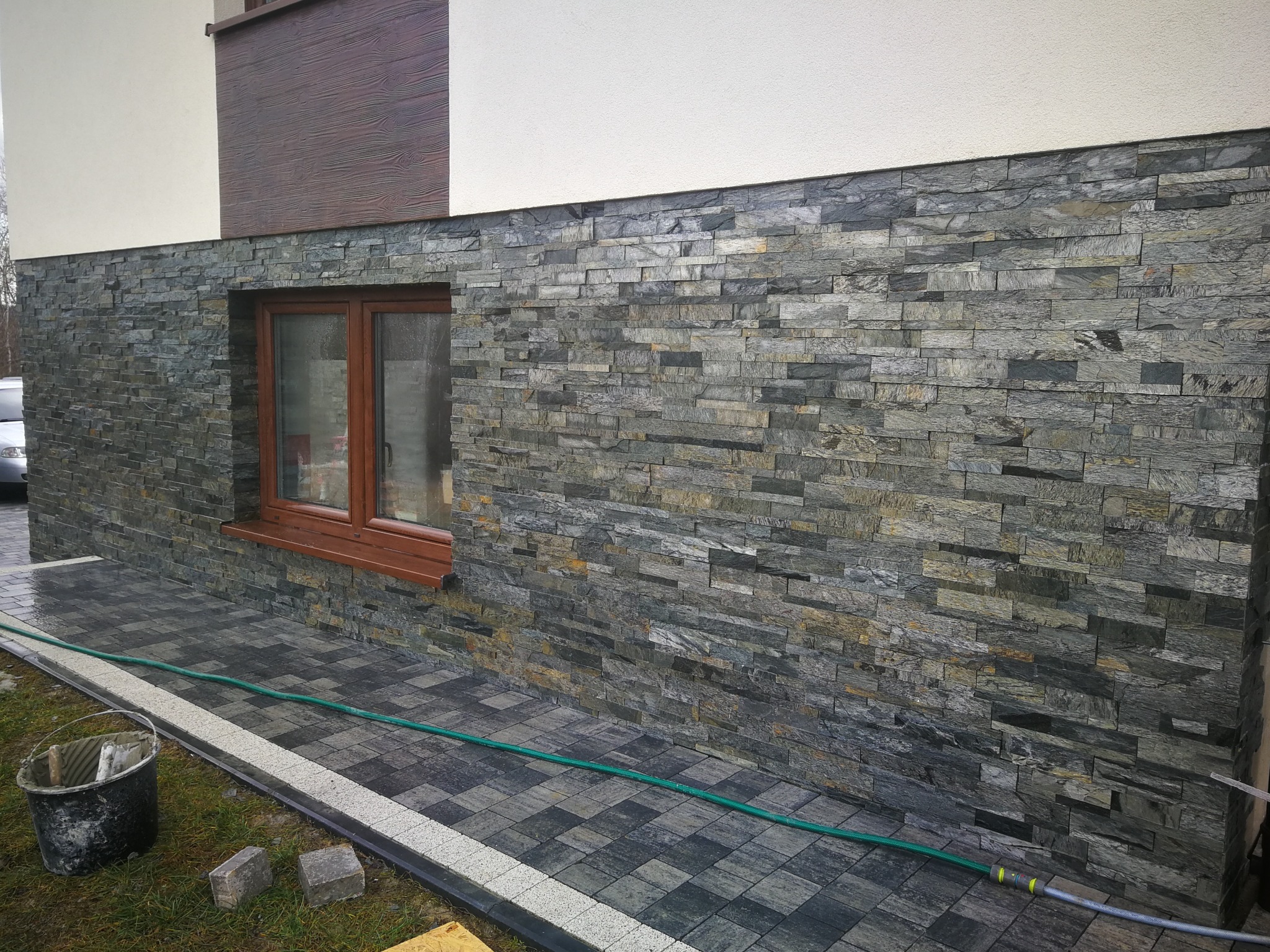 Elewacja budynku wykończona szarą, łupkową okładziną kamienną w wąskich pasach, z widocznym oknem z brązową ramą, dopasowana kolorystycznie do kostki brukowej i zarośniętym trawnikiem u podstawy.