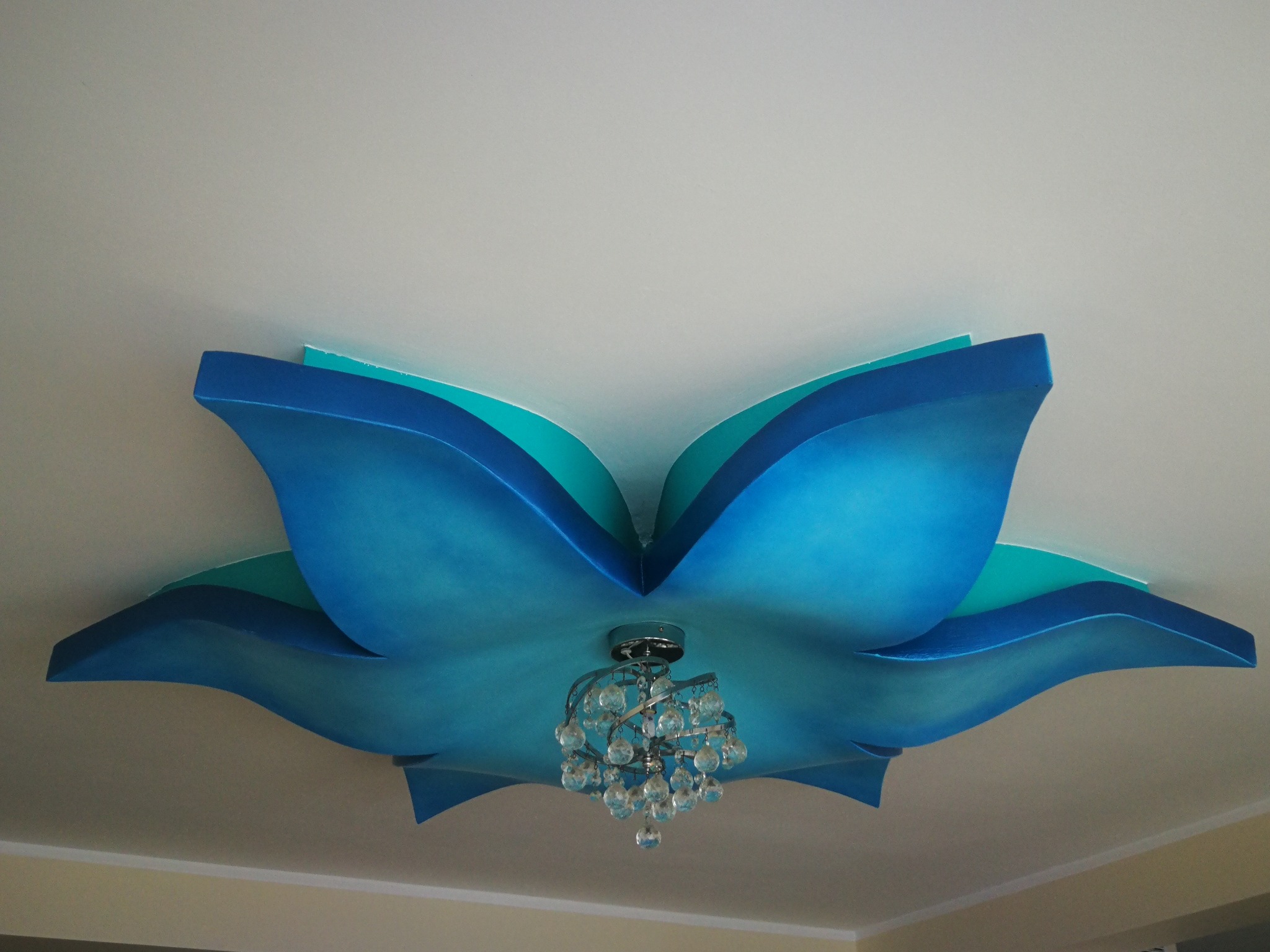 Dekoracyjny sufit w kształcie kwiatu w odcieniach błękitu i turkusu z zamontowanym żyrandolem z kryształkami.