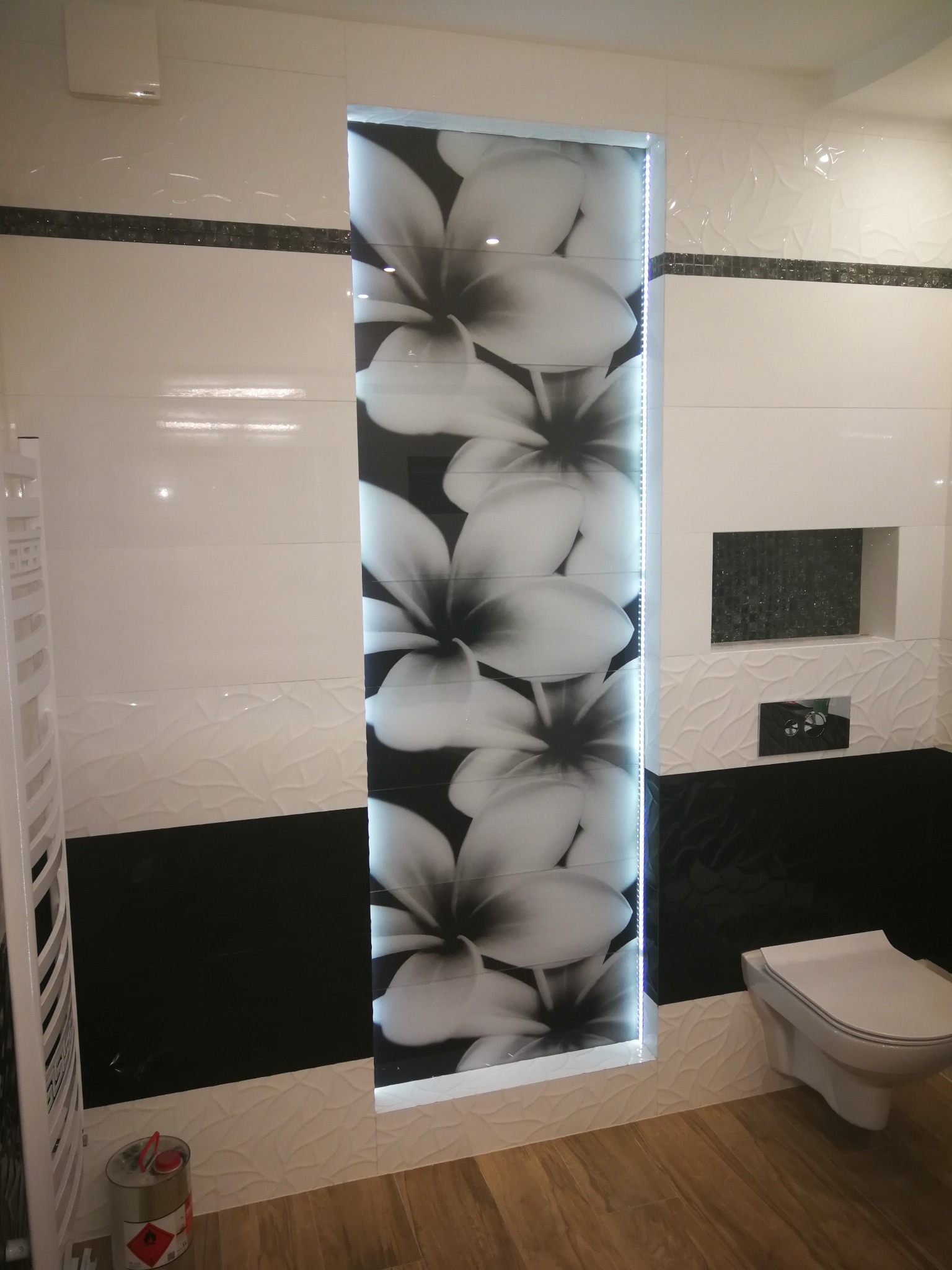 Wykończona łazienka z biało-czarnymi płytkami, podświetlaną wnęką z motywem kwiatowym, wiszącą toaletą i grzejnikiem drabinkowym, drewnopodobna podłoga.