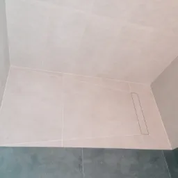 Wykończony brodzik prysznicowy z odpływem liniowym, wyłożony beżowymi i szarymi płytkami ceramicznymi o zróżnicowanej fakturze na ścianach i podłodze.