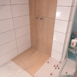 Układanie płytek imitujących drewno na ścianie i podłodze w łazience, z użyciem poziomujących klipsów i krzyżyków dystansowych, widoczne przyłącza wodne.