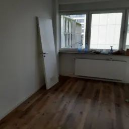 Pusty pokój z drewnianą podłogą, białymi ścianami i drzwiami, grzejnikiem pod oknem, przygotowany do prac wykończeniowych.