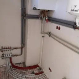 Instalacja ogrzewania podłogowego z rozdzielaczem, rurami w izolacji i kotłem gazowym na ścianie w surowym pomieszczeniu.