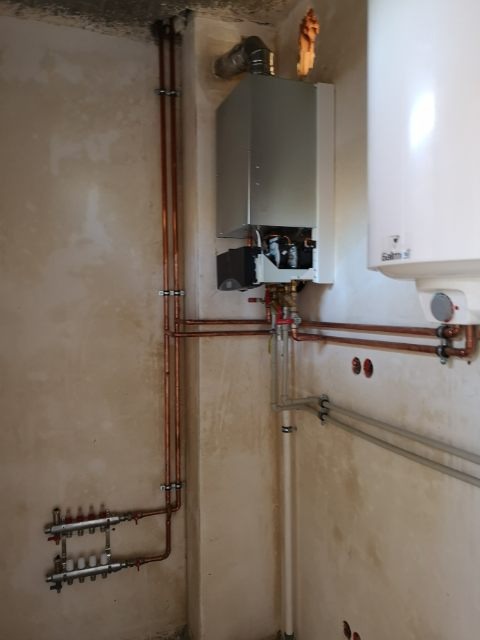 Nowy, wiszący piec gazowy z instalacją rur miedzianych i rozdzielaczem podłogowym zamontowany na surowej ścianie w narożniku pomieszczenia.