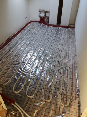Instalacja ogrzewania podłogowego: białe rurki ułożone w pętle na folii z czerwoną siatką, czerwone rury wzdłuż ścian, kolektor w tle.