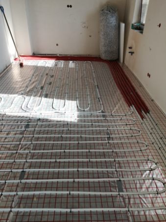 Instalacja ogrzewania podłogowego: białe rury ułożone na czerwonej siatce, częściowo przykryte folią, widoczne pionowe rury przy ścianie i worek z materiałem izolacyjnym.