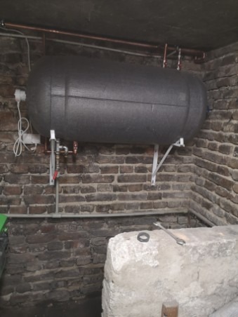 Zamontowany poziomy zbiornik akumulacyjny ciepłej wody w piwnicy z ceglanymi ścianami. Widoczne miedziane rury, białe wsporniki i przewód zasilający. Na pierwszym planie betonowa konstrukcja...