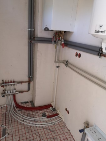 Instalacja ogrzewania podłogowego z rozdzielaczem, rurami w izolacji i kotłem gazowym na ścianie w surowym pomieszczeniu.