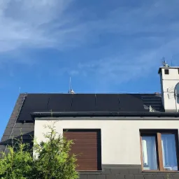 Panele słoneczne na dachu domu jednorodzinnego, widok z dołu na tle błękitnego nieba z antenami satelitarnymi na kominie.