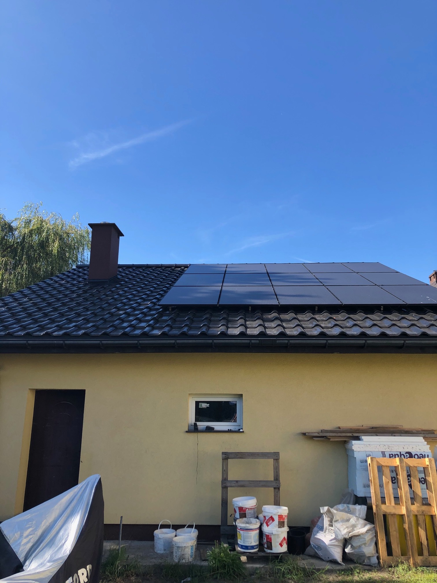 Dom z żółtą elewacją, na dachu częściowo zainstalowane panele słoneczne, widoczne materiały budowlane i puste wiadra po farbie przy ścianie budynku.