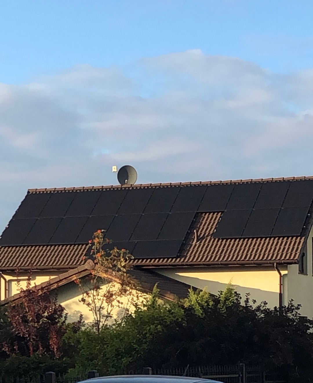 Dom jednorodzinny z panelami fotowoltaicznymi na dachu, widoczne z poziomu ulicy, z anteną satelitarną i fragmentem nieba w tle.