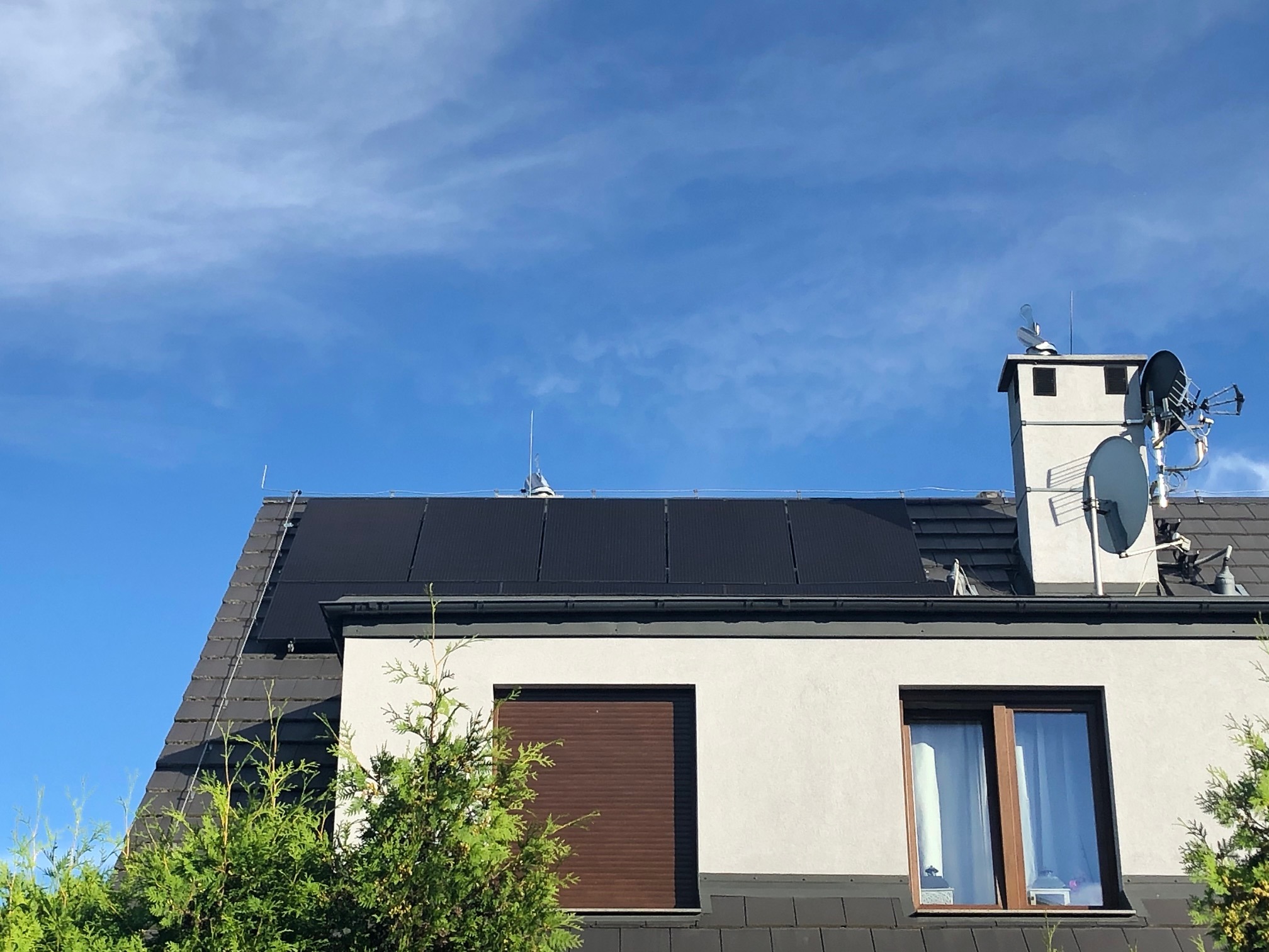 Panele słoneczne na dachu domu jednorodzinnego, widok z dołu na tle błękitnego nieba z antenami satelitarnymi na kominie.