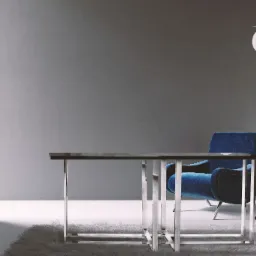 Nowoczesna konsola z czarnym blatem i chromowanymi nogami na tle szarej ściany, obok niebieski fotel i lampa podłogowa z kulistym kloszem.