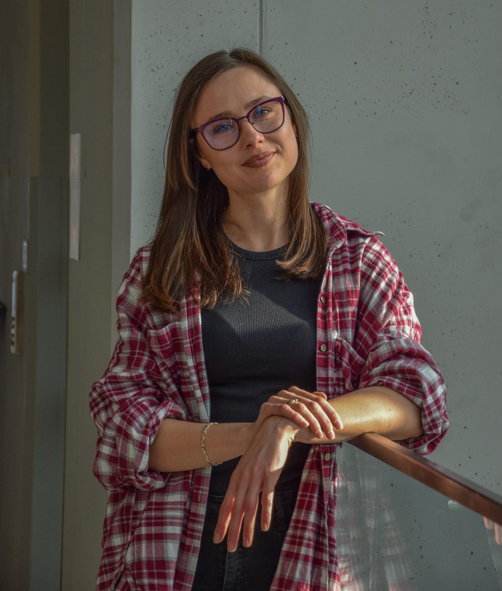 Kobieta w okularach i flanelowej koszuli opiera się o szklaną barierkę na tle betonowej ściany w nowoczesnym wnętrzu.