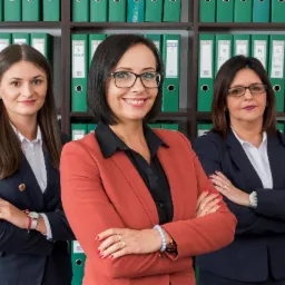 Zespół trzech kobiet w biurze, uśmiechnięte, w eleganckich strojach na tle regału z segregatorami z dokumentami.