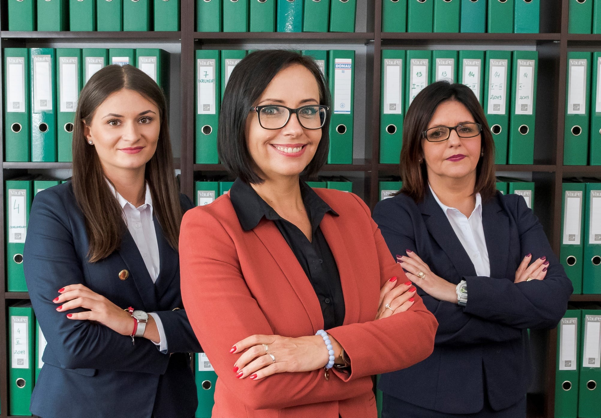 Zespół trzech kobiet w biurze, uśmiechnięte, w eleganckich strojach na tle regału z segregatorami z dokumentami.